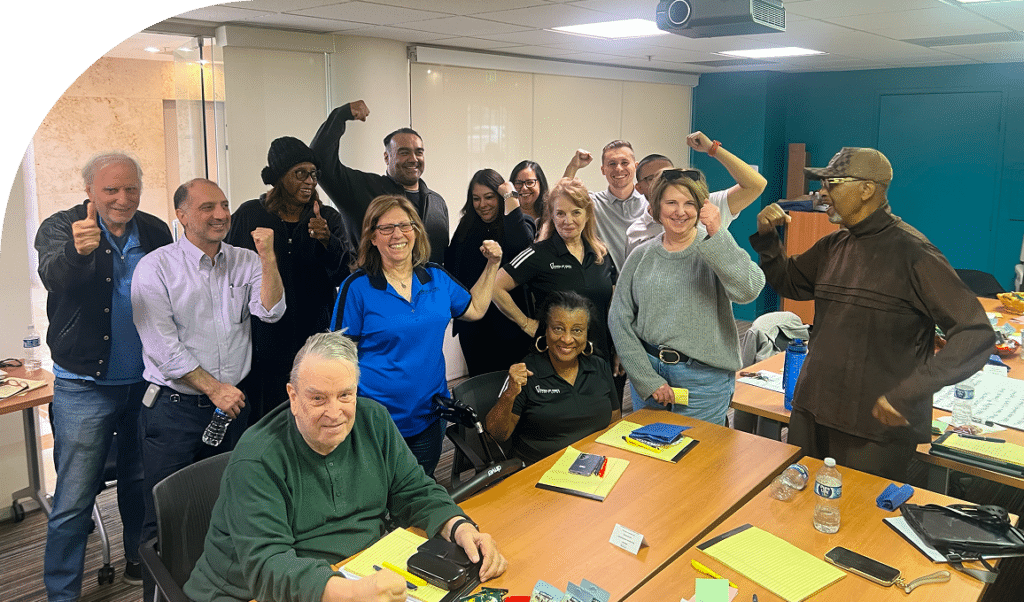 A group of thirteen people in an office setting, displaying cheerful expressions and victory poses. Two people sit at a wooden table; others stand behind them.