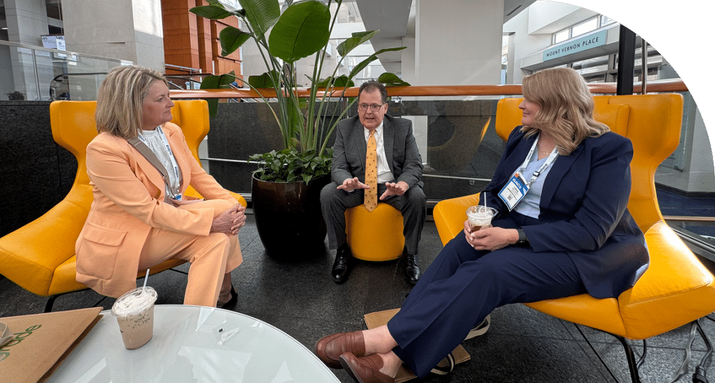Three people sit in vibrant yellow chairs around a small table, engaging in conversation. They appear relaxed, with a plant in the background and drinks in hand.