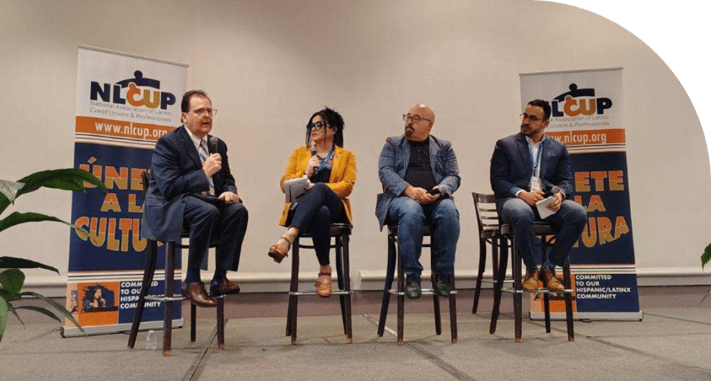 Four people are seated on high chairs in a panel discussion setting. Two banners with "NLCUP" and "Únete a la Cultura" flank the speakers, conveying a professional atmosphere.