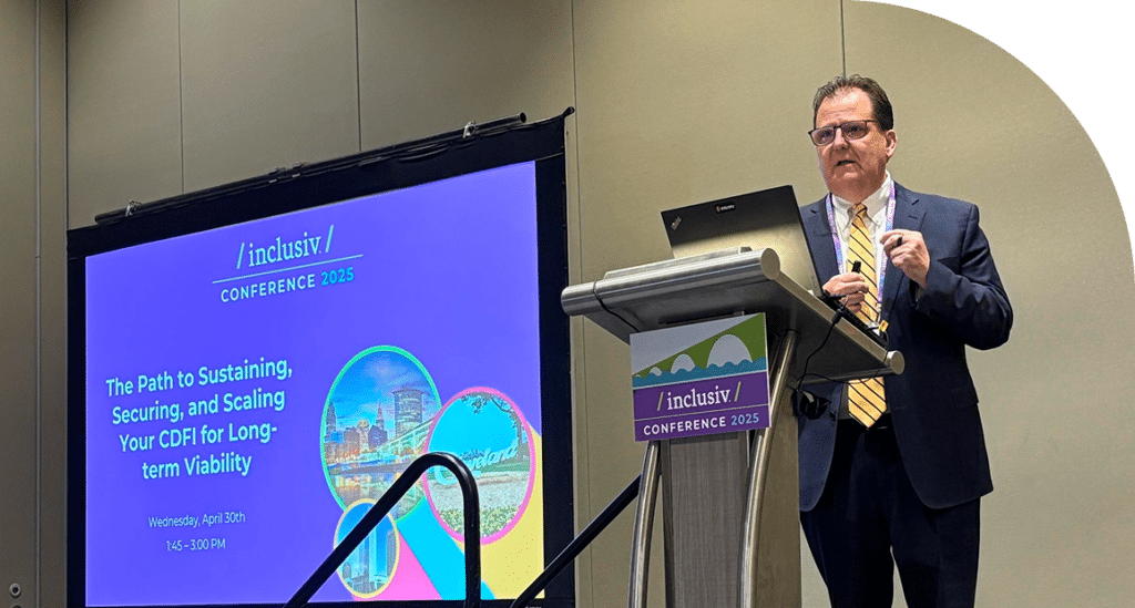A speaker in a suit presents at a podium with a laptop at Inclusiv Conference 2025. A screen displays conference details and colorful graphics.