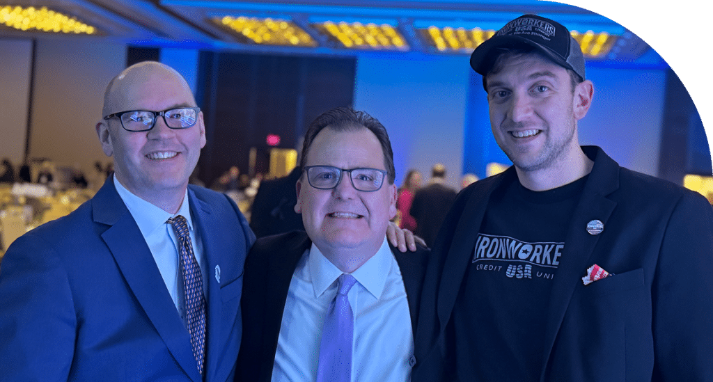 Three smiling men stand closely together at an indoor event with a blue-lit background. Two wear suits and ties, one in a cap and casual jacket.