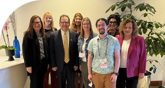 Eight people, smiling, stand indoors in business attire. A man in a suit is in front, surrounded by others in blazers. A plant is in the background.