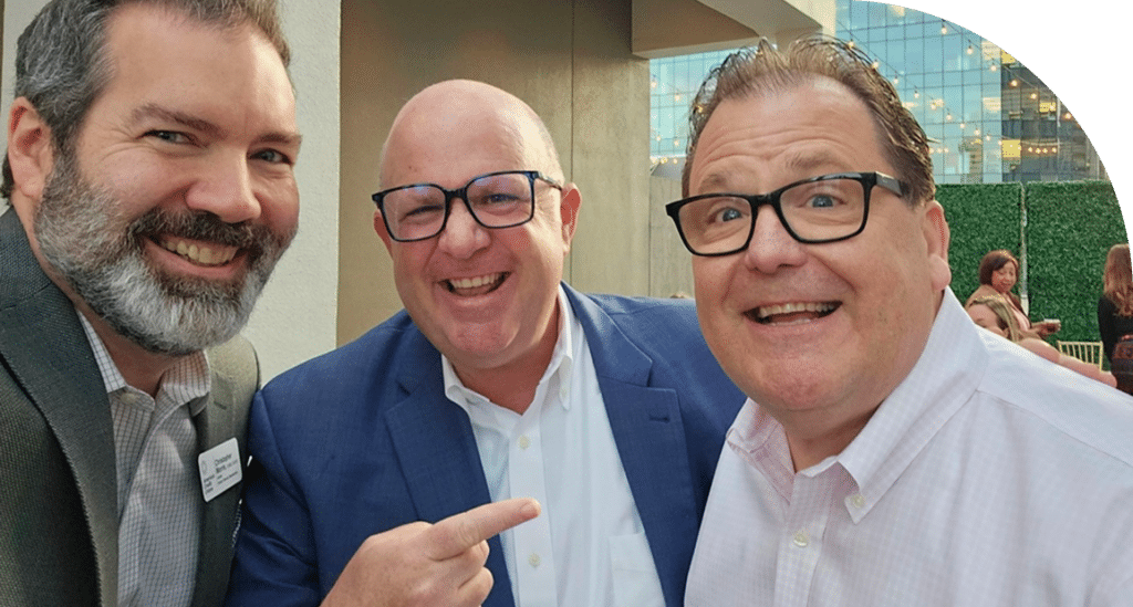 Three men, smiling joyfully, take a selfie at an outdoor event. The background shows modern buildings and greenery, suggesting a lively atmosphere.