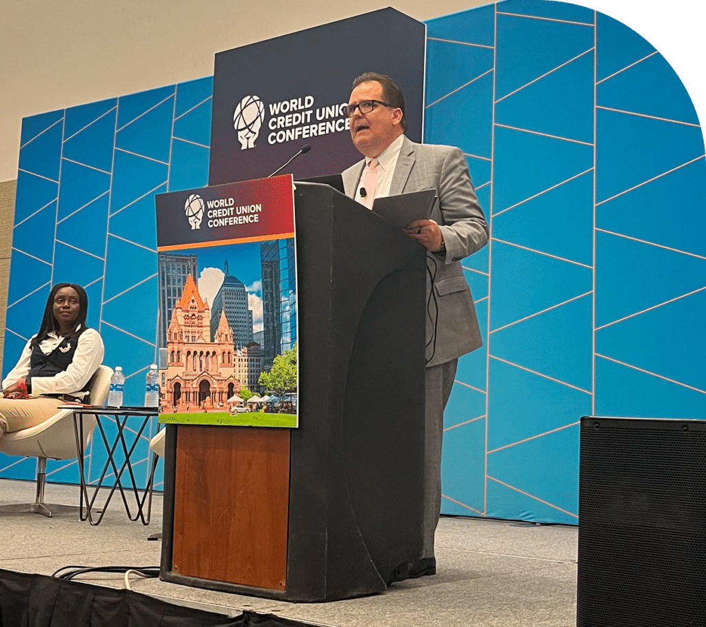 A man in a gray suit speaks at a podium during the World Credit Union Conference with a cityscape backdrop. A woman sits to the left, listening attentively.