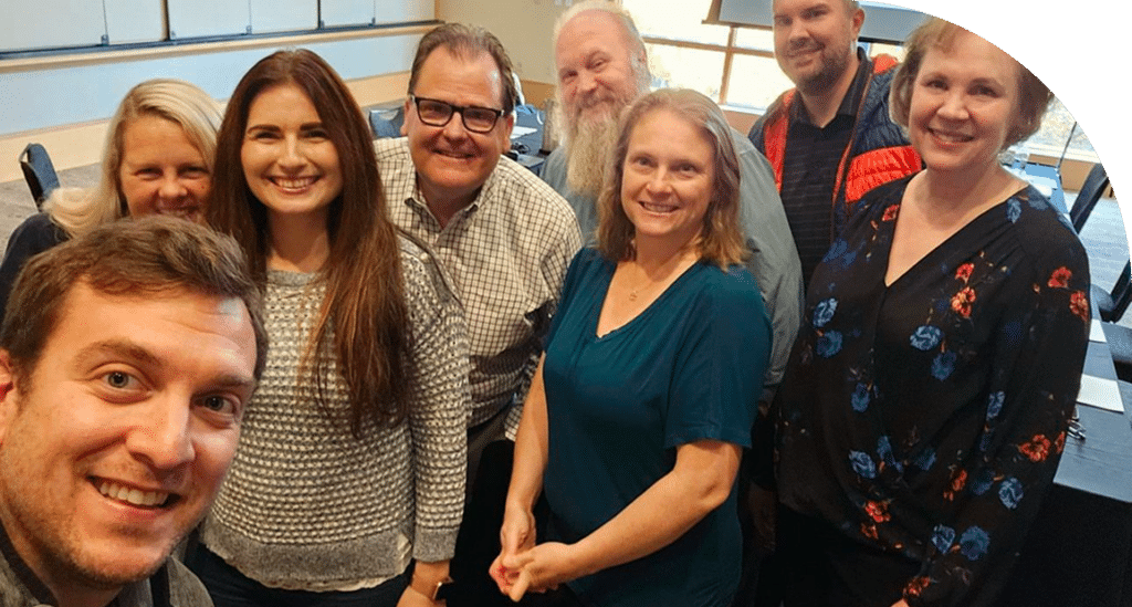 A group of eight smiling people pose for a selfie in a conference room. They appear friendly and relaxed, conveying a cheerful and welcoming atmosphere.