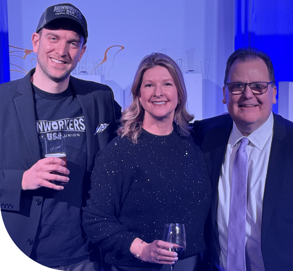 Three people smiling at an event. The man on the left wears a cap and holds a beer, the woman in the middle has a sparkly sweater, and the man on the right wears glasses and a tie. The atmosphere is warm and convivial.