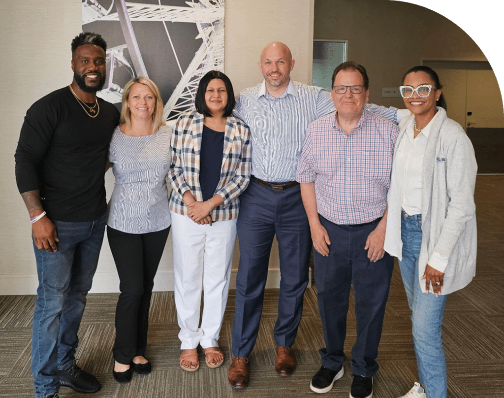 A diverse group of six people stand closely together, smiling in a professional setting. They exude a welcoming, collaborative atmosphere.