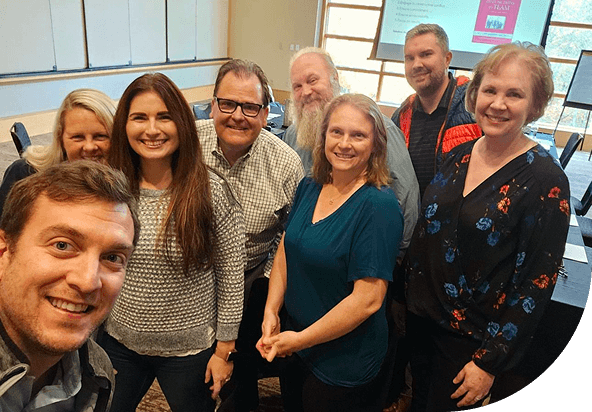 A group of eight smiling people pose for a selfie in a conference room. They appear friendly and relaxed, conveying a cheerful and welcoming atmosphere.