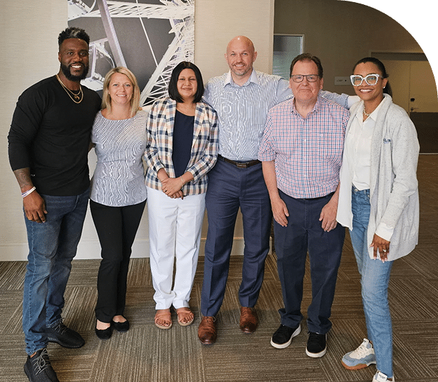 A diverse group of six people stand closely together, smiling in a professional setting. They exude a welcoming, collaborative atmosphere.