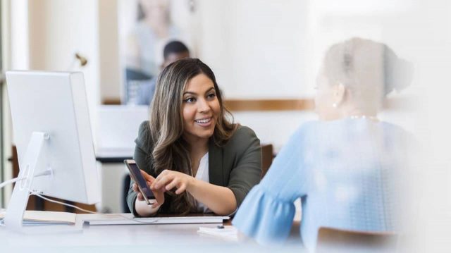 Woman sitting at desk holding cell phone sharing screen with woman on other side of desk.